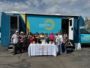 Group of people holding flowers in front of a blue Marisol Health bus, standing by a table with promotional materials. Sunny day.