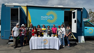 Group of people holding flowers in front of a blue Marisol Health bus, standing by a table with promotional materials. Sunny day.