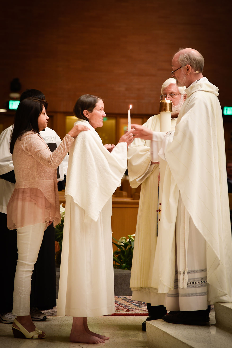 A solemn ceremony with a woman holding a candle, draped in a white robe, assisted by others in a church. Soft lighting, peaceful mood.