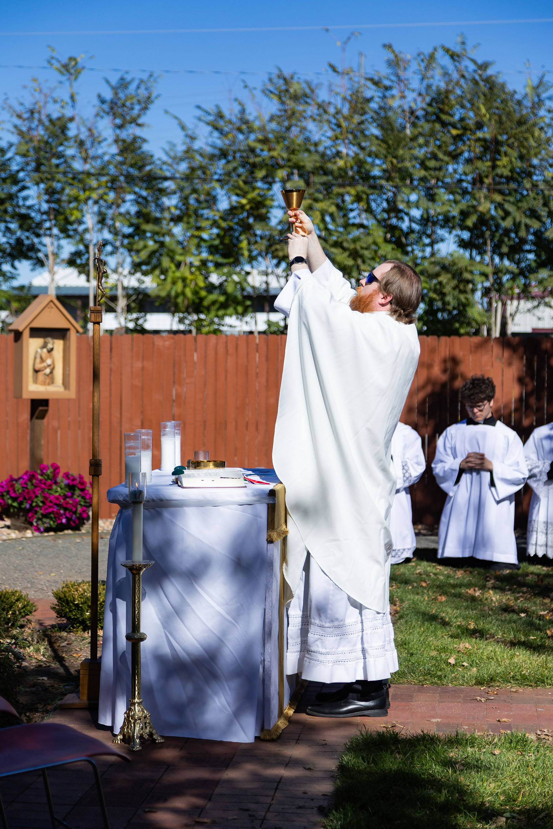 A priest in white robes holds a chalice aloft outdoors beside an altar, with acolytes in white robes nearby. A red fence and trees are in the background.