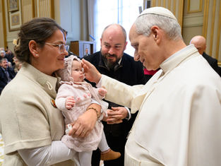 Clergy member blesses a baby held by a woman. Man stands beside them. Ornate room with gold accents. Warm, serene atmosphere.