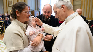 Clergy member blesses a baby held by a woman. Man stands beside them. Ornate room with gold accents. Warm, serene atmosphere.