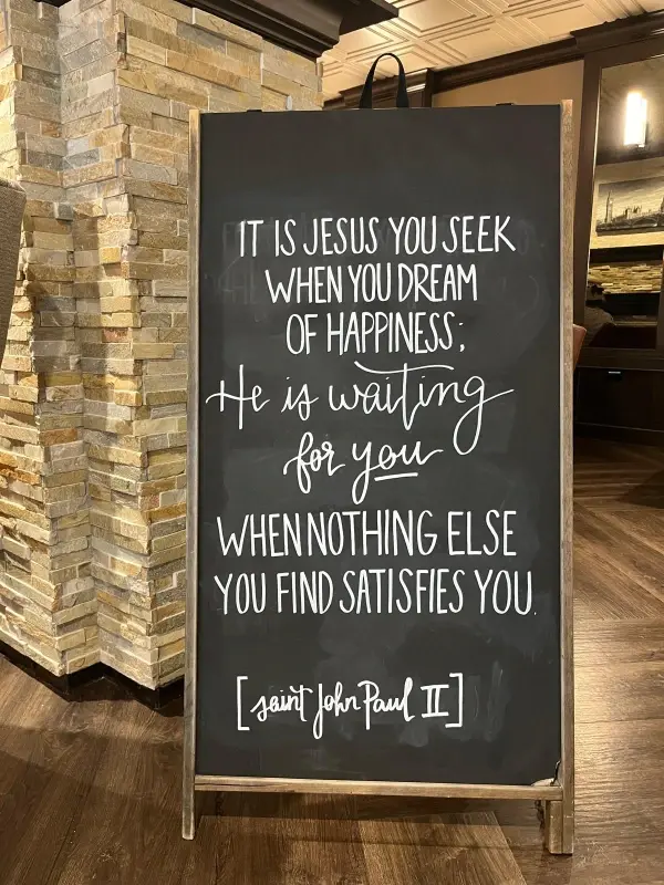 Chalkboard with message about seeking Jesus for happiness, attributed to Saint John Paul II. Set in an indoor space with stone walls.