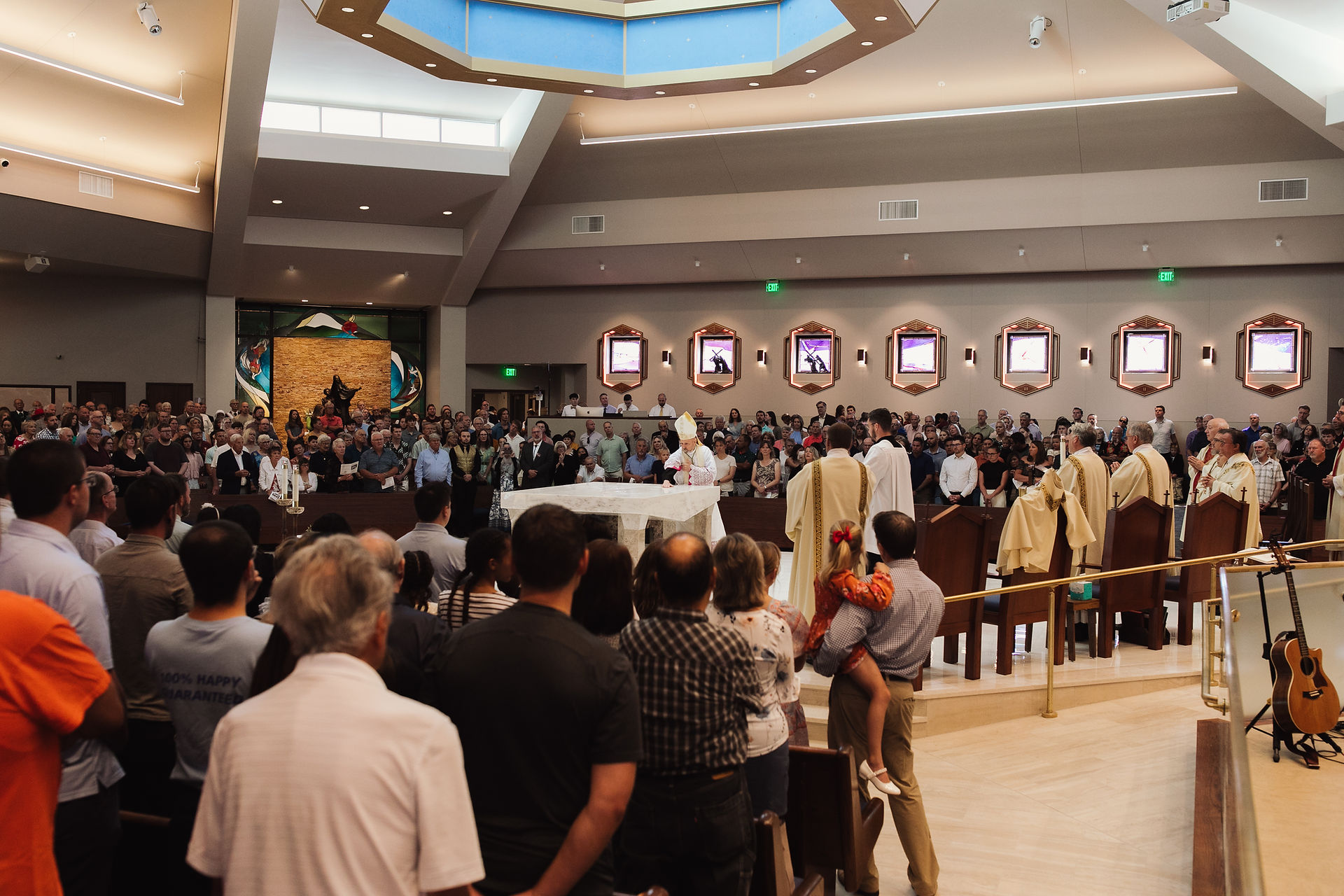 A large congregation gathers in a modern church, focusing on a priest near the altar. Warm lighting, decorative paintings, and a guitar are visible.
