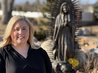 Woman in black shirt smiling outdoors near a religious statue with flowers. Sunny day, natural setting, and peaceful mood.