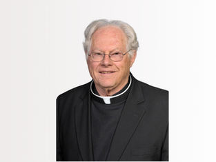 Portrait of an elderly priest in black clerical attire and glasses, smiling against a plain white background.