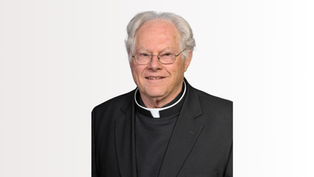 Portrait of an elderly priest in black clerical attire and glasses, smiling against a plain white background.