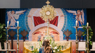 A person kneels facing a ornate monstrance on an altar with candles. Religious mural backdrop features angels, rich colors, and patterns.