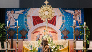 A person kneels facing a ornate monstrance on an altar with candles. Religious mural backdrop features angels, rich colors, and patterns.