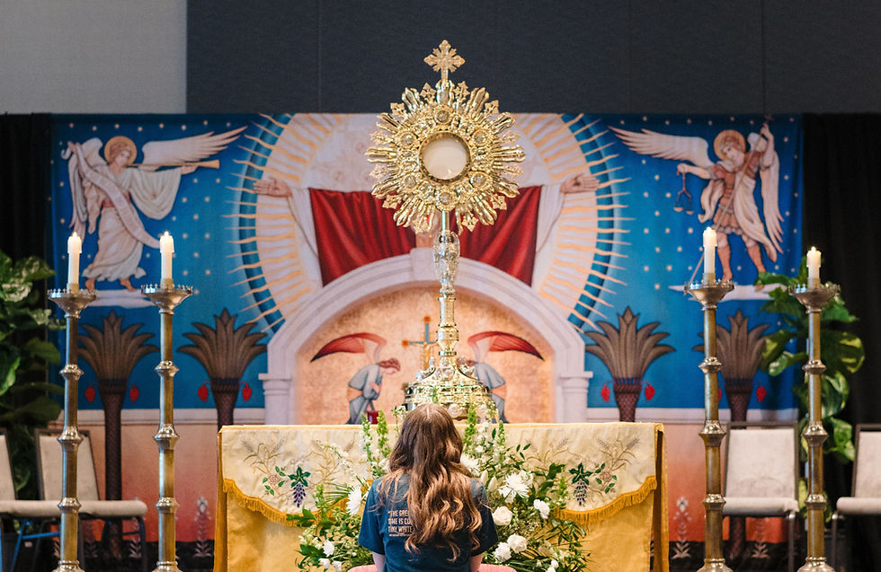 A person kneels facing a ornate monstrance on an altar with candles. Religious mural backdrop features angels, rich colors, and patterns.