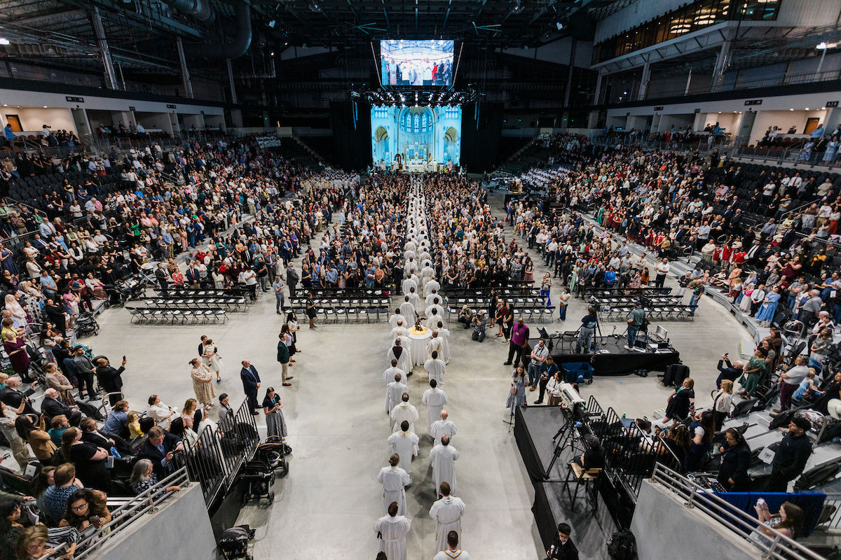 A large procession of clergy moves down the center aisle of a packed arena during a Catholic installation Mass.