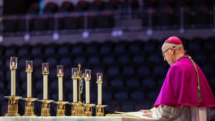 A bishop in purple reads at an altar with lit candles and a crucifix. The background is a dimly lit auditorium, creating a solemn mood.