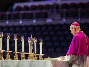 A bishop in purple reads at an altar with lit candles and a crucifix. The background is a dimly lit auditorium, creating a solemn mood.