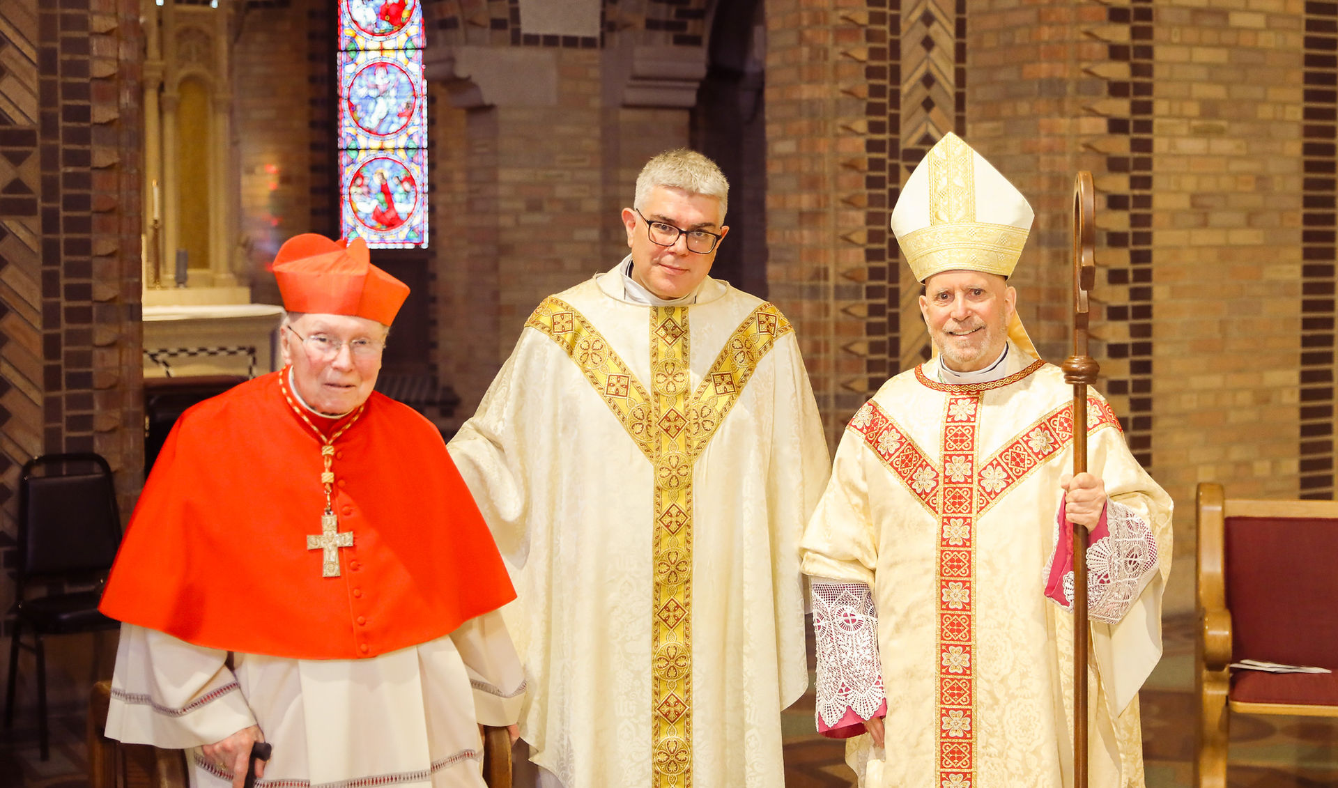 A cardinal, a priest and an archbishop stand, smiling for a photo, in an ornate brick chapel following a religious ceremony.