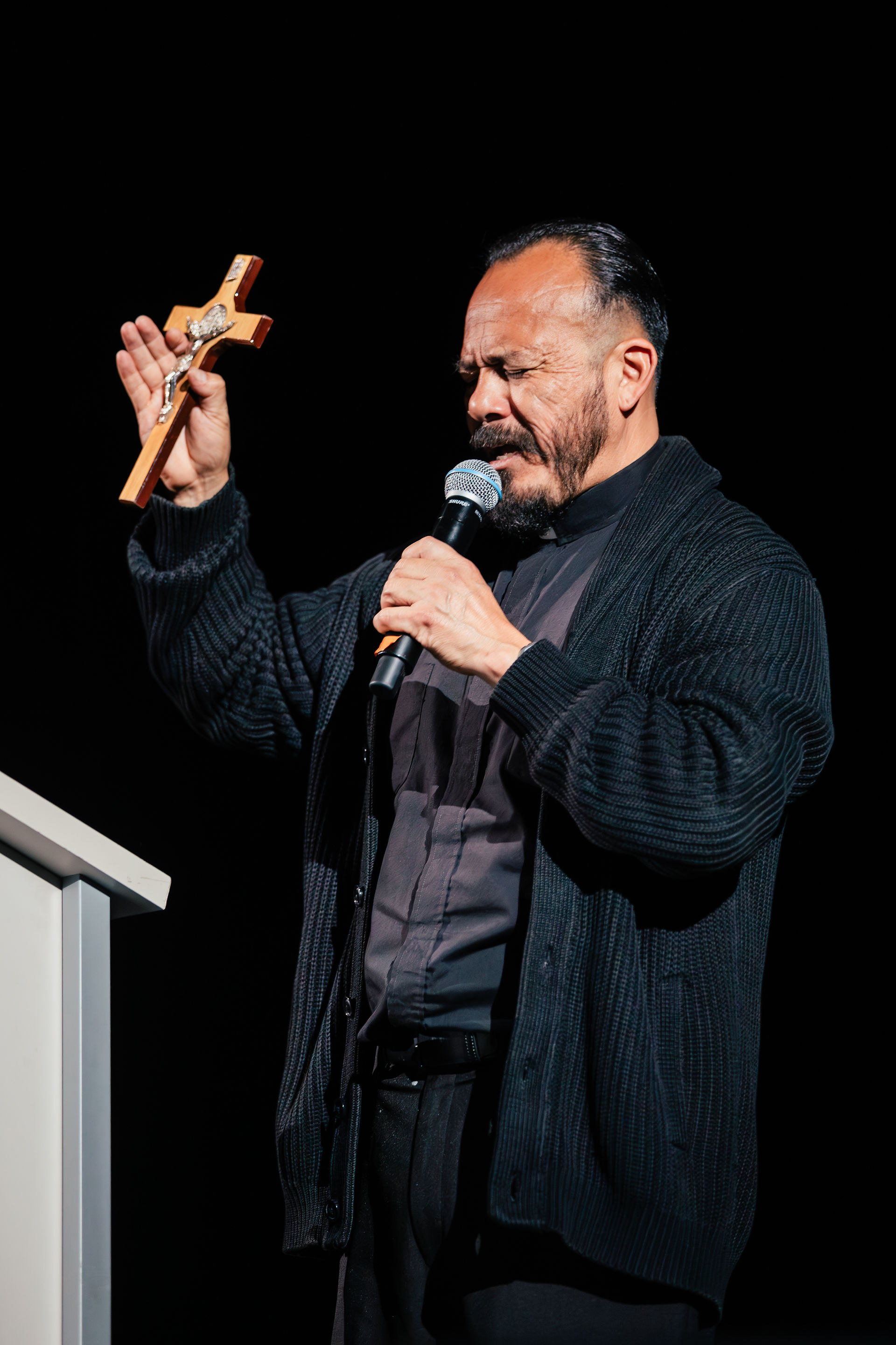 Man in a black sweater holding a cross and microphone, eyes closed in focus, standing at a podium against a dark background.