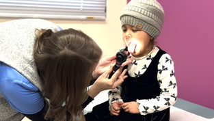 Child in a hat getting throat examined by a woman with an otoscope. Bright room with purple wall, calm atmosphere.