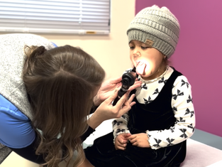 Child in a hat getting throat examined by a woman with an otoscope. Bright room with purple wall, calm atmosphere.