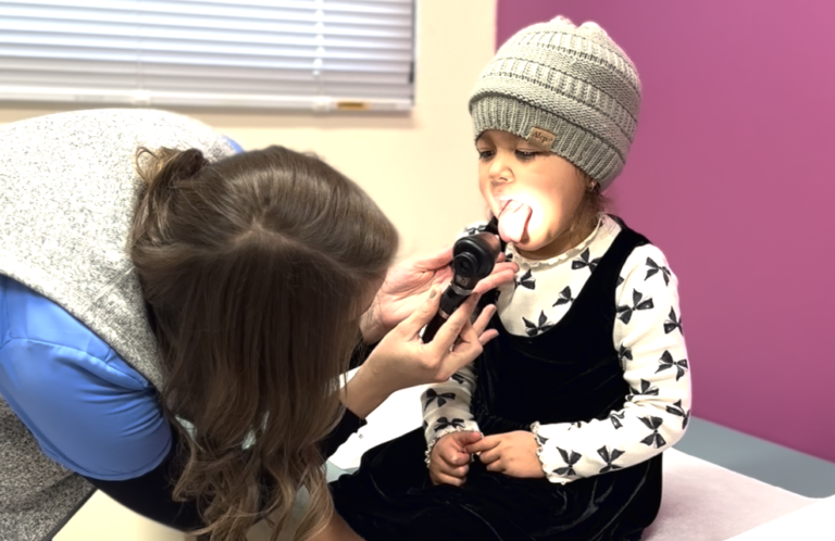 Child in a hat getting throat examined by a woman with an otoscope. Bright room with purple wall, calm atmosphere.