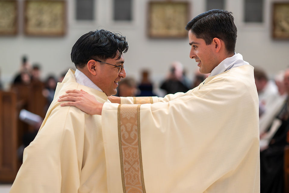 Two deacons in cream robes smile warmly at each other, one placing hands on the other's shoulders. Blurred church background, joyful mood.