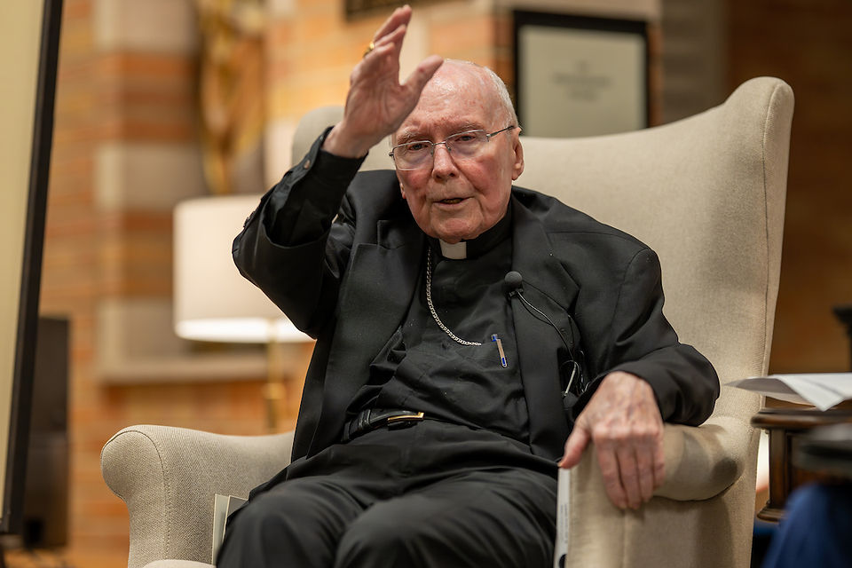 Elderly man in black clerical attire sits in an armchair, raising his hand in. blessing. Warm interior with books in the background; focused expression.