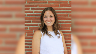 Smiling woman in a white top stands against a brick wall. She's looking at the camera, creating a cheerful mood.