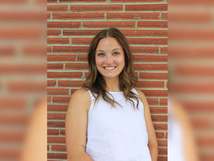 Smiling woman in a white top stands against a brick wall. She's looking at the camera, creating a cheerful mood.