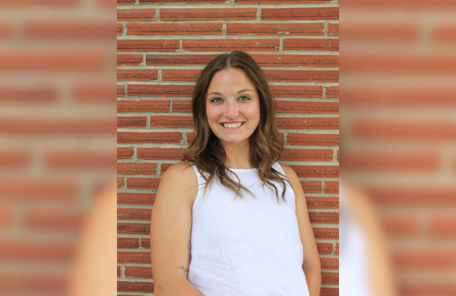 Smiling woman in a white top stands against a brick wall. She's looking at the camera, creating a cheerful mood.