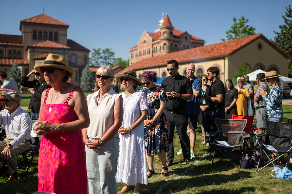 People stand in a sunlit line on a grassy lawn, some in hats and sunglasses, at an outdoor ceremony by red-tiled buildings.