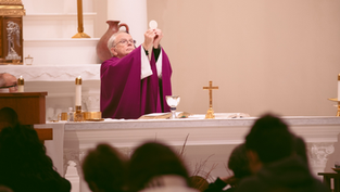 Priest in purple vestments holds up the Eucharist in a church. Altar with candles, chalice, and cross. Congregation in foreground.