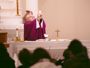 Priest in purple vestments holds up the Eucharist in a church. Altar with candles, chalice, and cross. Congregation in foreground.