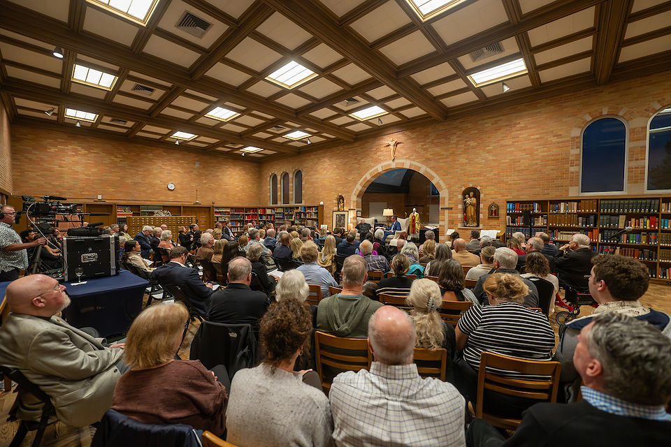 Audience in a library with brick walls and statues listens to a speaker. Camera crew on the left. Warm lighting, high ceiling.