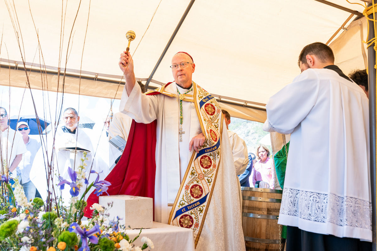 Bishop in white and red vestments raises a staff at an outdoor Mass under a tent, with flowers and worshippers nearby