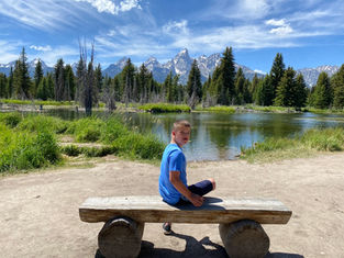 Boy in a blue shirt sits on a wooden bench by a lake, under a bright sky. Mountains and pine trees form a scenic backdrop.