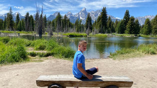Boy in a blue shirt sits on a wooden bench by a lake, under a bright sky. Mountains and pine trees form a scenic backdrop.