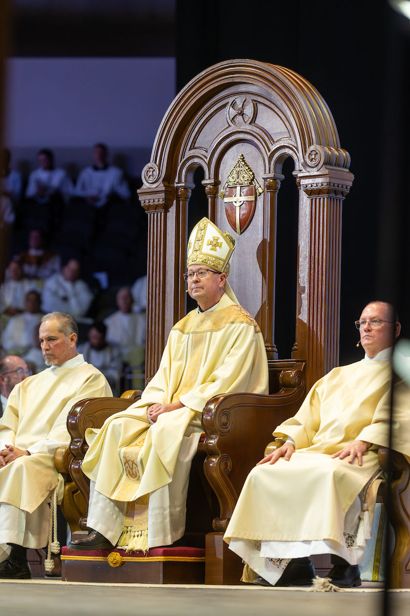 A bishop in ornate robes and mitre sits on a grand wooden chair, flanked by two priests in a ceremonial setting, with onlookers blurred in the background.