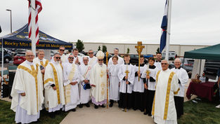 Clergy and altar servers in white and gold robes pose outdoors by a flagpole. "Rocky Mtn Regional VA Medical Center" visible on a canopy.