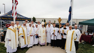Clergy and altar servers in white and gold robes pose outdoors by a flagpole. "Rocky Mtn Regional VA Medical Center" visible on a canopy.