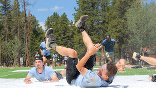 People tumble on a wet outdoor slip-n-slide, splashing on grass under pine trees in a sunny park.