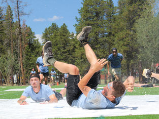 People tumble on a wet outdoor slip-n-slide, splashing on grass under pine trees in a sunny park.