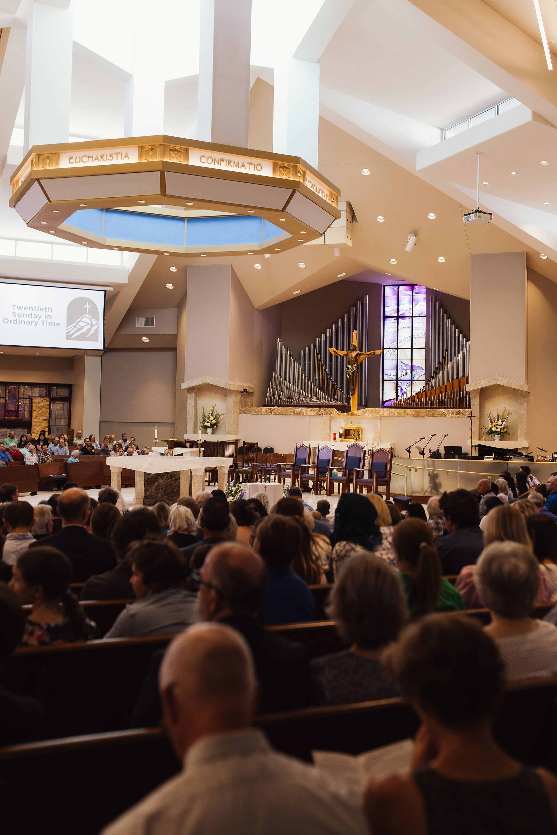 Congregation seated in a modern church interior during a service. Large organ, crucifix, and text "Twentieth Sunday in Ordinary Time."