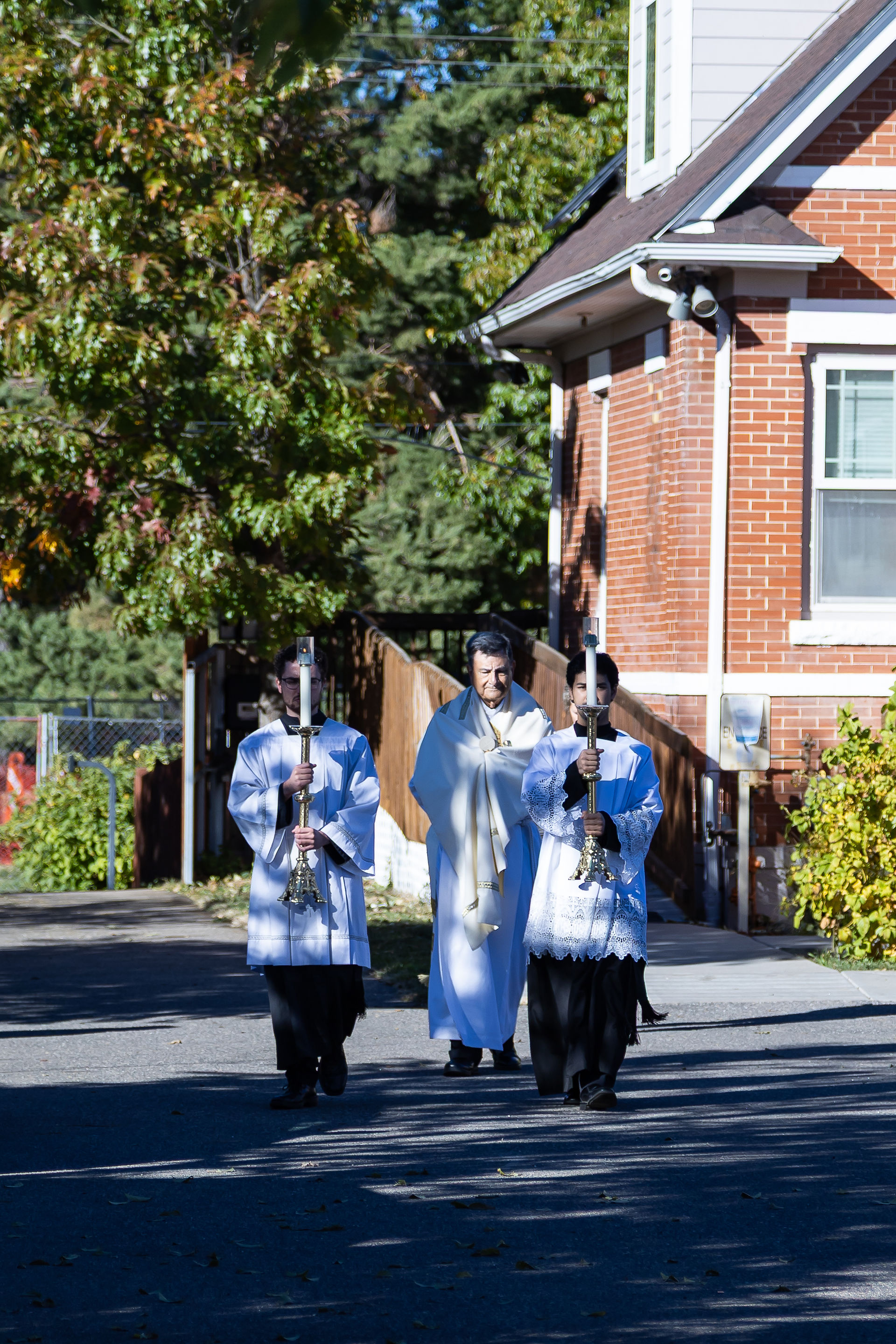 Three people in white robes walk outside a brick building, holding candles. Sunny day with green trees in the background.