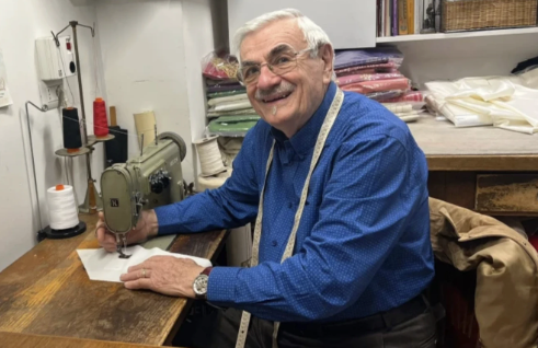 Older man in a blue shirt smiles while sewing at a machine. Shelves with colorful fabrics are in the background, creating a cozy atmosphere.