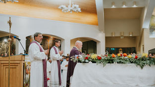 Clergy in white robes and purple stoles stand and pray by a flower-covered altar in a church. The wooden ceiling and a cross are visible.