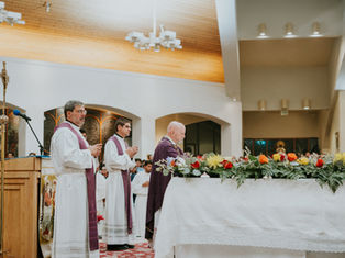 Clergy in white robes and purple stoles stand and pray by a flower-covered altar in a church. The wooden ceiling and a cross are visible.