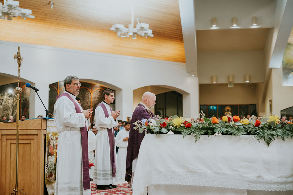 Clergy in white robes and purple stoles stand and pray by a flower-covered altar in a church. The wooden ceiling and a cross are visible.