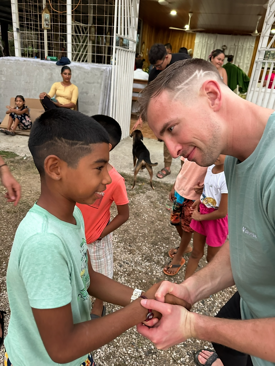 Man and boy smiling and shaking hands outdoors. Other people and a dog in the background. Casual setting with warm, friendly mood.