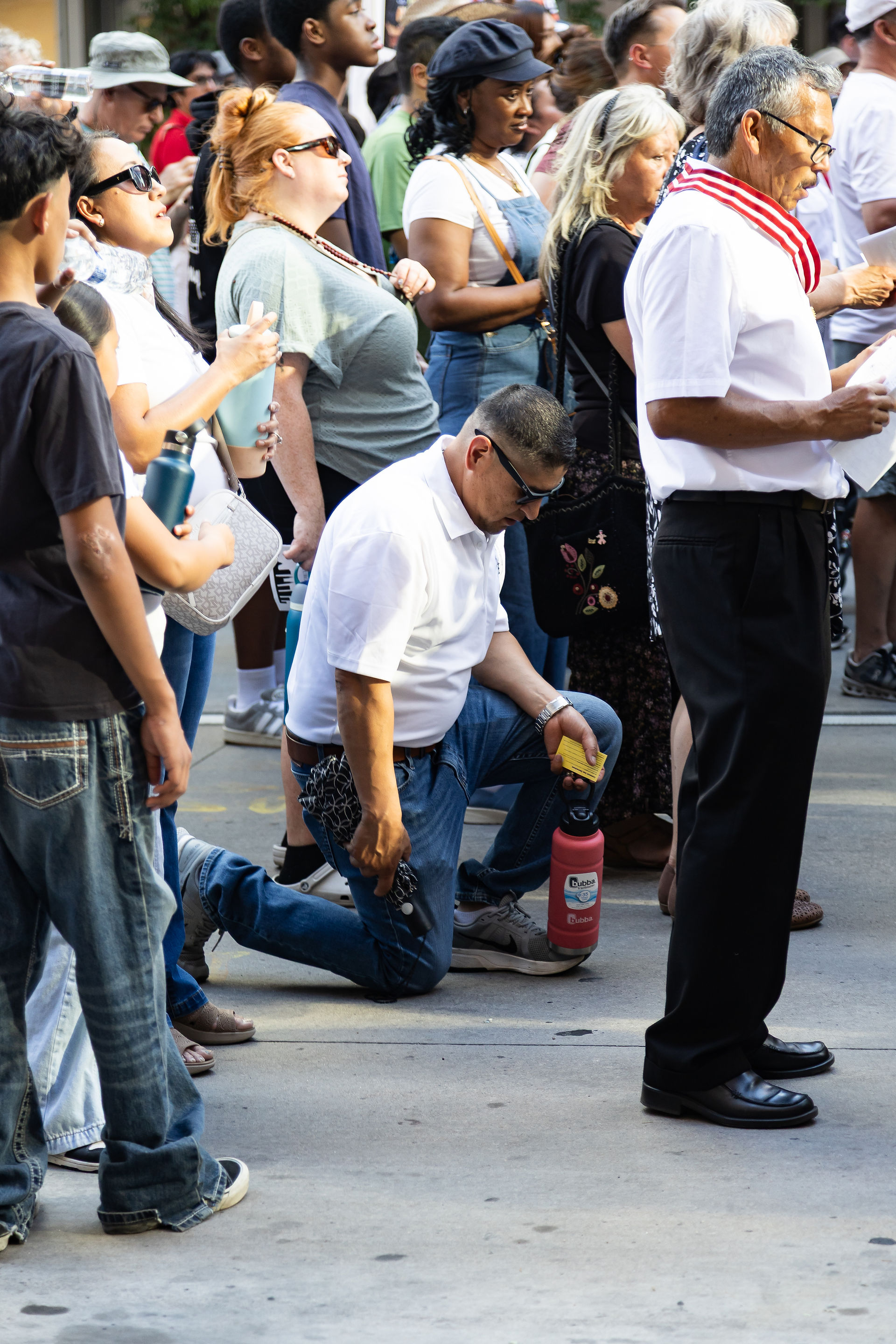 Crowd at an outdoor gathering, with a man kneeling in a white shirt and others standing solemnly, holding water bottles.