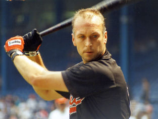 Baseball player in black jersey and orange gloves poised to swing a bat. Focused expression. Stadium background with blurred audience.