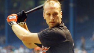 Baseball player in black jersey and orange gloves poised to swing a bat. Focused expression. Stadium background with blurred audience.