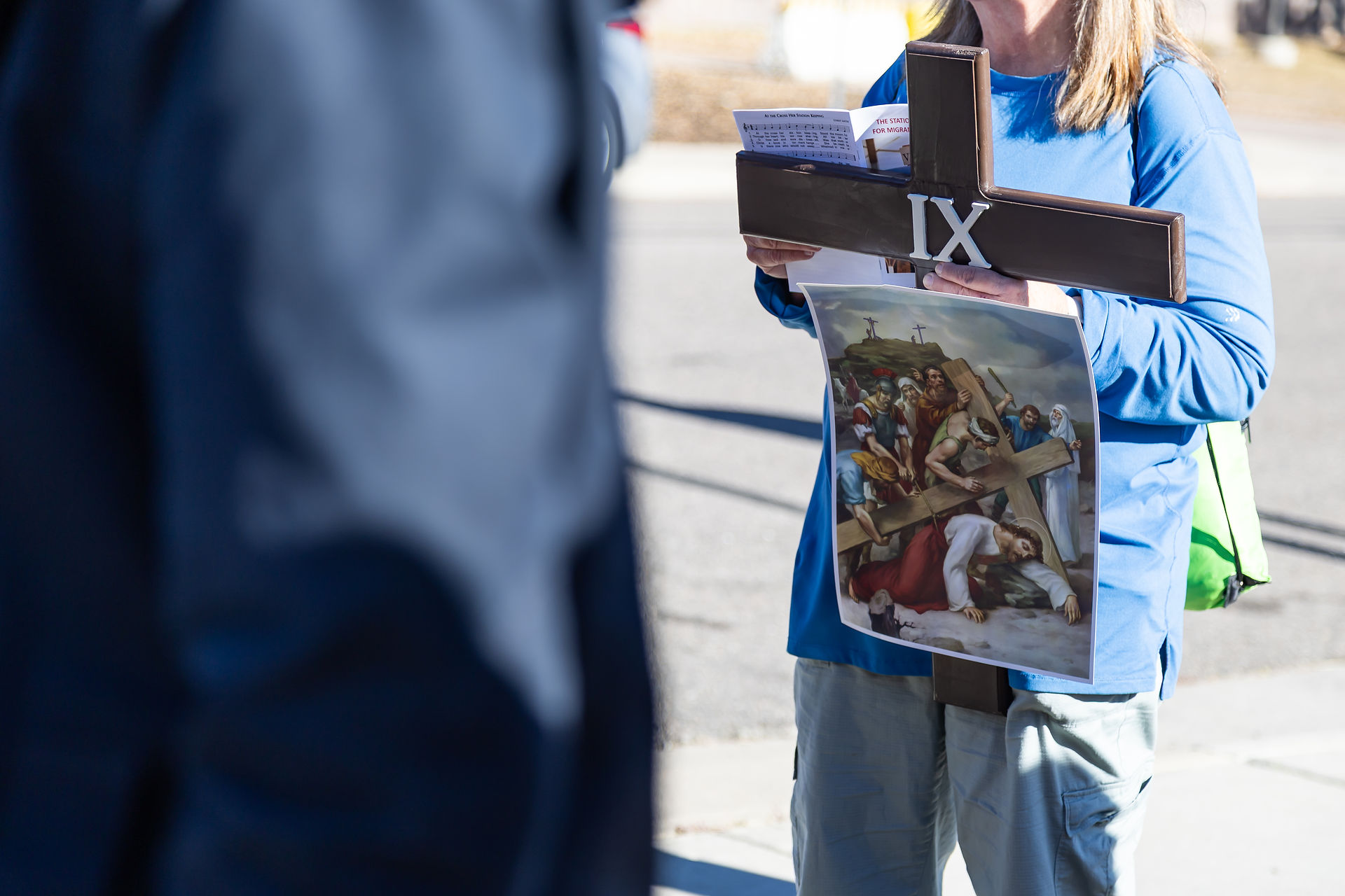 Person in blue holds a cross marked IX and a painting of Jesus carrying the cross. Background is a sunny street with a blurred figure.